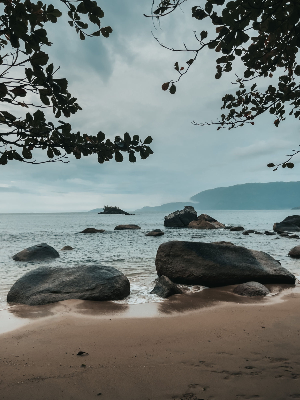Praia com pedras e mar calmo vistos entre galhos de árvore, em Ilhabela, litoral norte de São Paulo